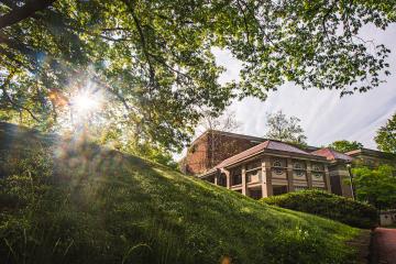 The sun rises behind a lush green hill and tree, with the Scripps Hall in the background