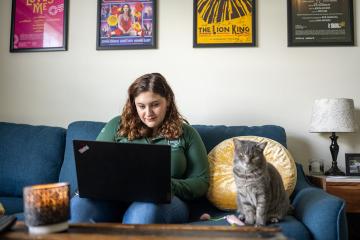 Woman in OHIO pullover works on a laptop while sitting on her sofa next to a gray tabby cat