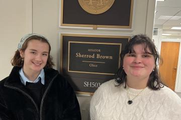 Two college students pose for a picture standing on either side of a sign that says "Senator Sherrod Brown"