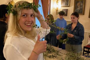 A student wearing a laurel wreath on her head smiles at the camera while others create wreaths in the background
