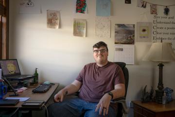 Christopher Johnson sits at the desk in his office with homemade art strung on the wall behind him