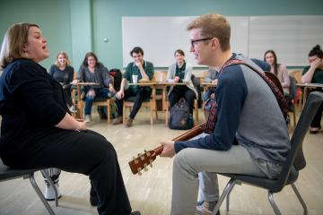 In a classroom, a student holds a guitar while seated across from the instructor as the class observes.