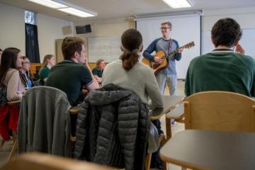 A man stands in front of a classroom full of students and plays the guitar