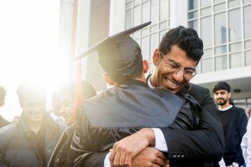 Two men, one wearing a graduation cap and gown, embrace as the sun shines behind them