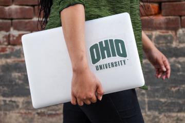 A person walking in front of a brick wall holding a laptop with an Ohio University sticker.