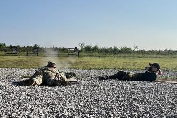 On a gravel surface, a soldier lying on the ground shoots at a target in the near distance while a photographer lies beside him, taking a picture