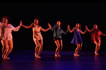 Five dramatically-lit dancers stand in a row, hands held up in connection with each other