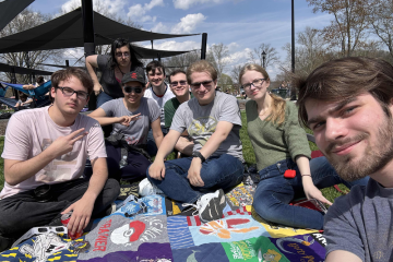 A group of people pose for a selfie, seated on a picnic blanket