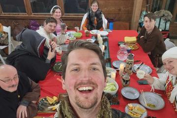 A man dressed in Medieval garb takes a selfie as a similarly dressed group sits behind him at a dining table