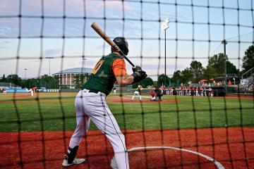 a Copperheads baseball player holds the bat as he waits on deck, viewed from behind a black safety net