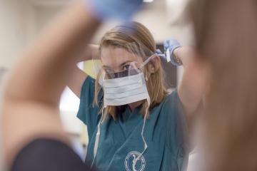 a female student wearing scrubs puts on a disposable face mask and plastic eye shield