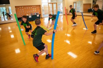 : two young boys and two older camp counselors play with pool noodles in a gymnasium-style room