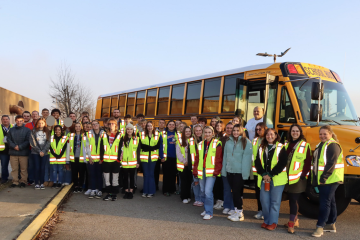 High school students from the region are show wearing safety vests while standing outside of a school bus