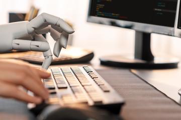 A robot hand typing on computer keyboard next to a human hand.