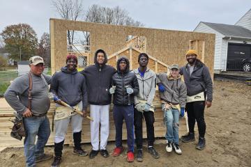 A group of men stands in front of a house mid-construction