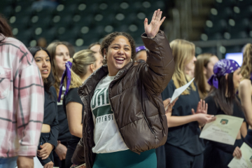 An OHIO student is shown waving to a friend while surrounded by other students in the Convocation Center