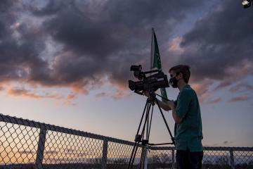 Bryan Kurp stands behind a camera wearing a mask, with a dramatic sunset as background