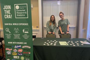 two young women stand behind a table encouraging students to sign up for the Center for Research and Analytics