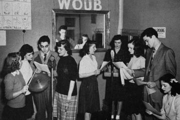 black-and-white photo of a group of students crowded around two microphones holding scripts in an early WOUB studio