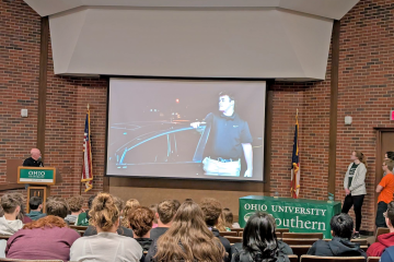 Students in auditorium look toward screen, man stands at podium