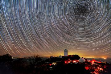  A long-exposure image captures circular star trails above telescope domes illuminated in red light on a dark mountain. A golden glow on the horizon marks distant city lights.