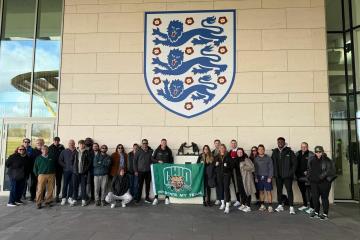 A group poses in front of the England football crest, holding an Ohio University flag outside a modern sports facility.