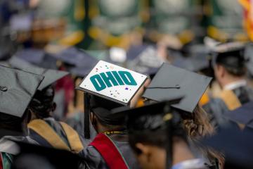 An OHIO student wears a commencement cap that says "OHIO" while sitting in a large group of students during the Commencement ceremony in the Convocation Center