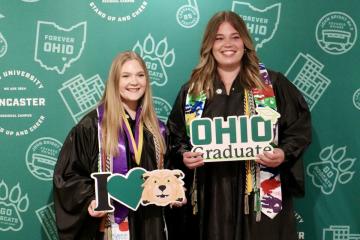 Two OHIO Lancaster graduates are shown in their graduation robes while holding OHIO Graduate signs