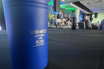A sign that says TechGrowth partnering with AOIA Athens OHIO Investment Alliance is shown at a media event where speakers are sitting on a stage