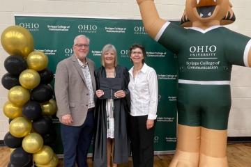 Mary Stephan is shown in commencement robe, holding her cap, standing with Scott Titsworth and Ann Bainbridge while they stand next to balloons and a blow-up figure of Rufus wearing a Scripps College of Communication shirt