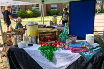 A Welcome Week  table is set up outside of an OHIO residence hall with apples and snacks and other items for families moving students into the residence halls. Volunteers are shown working at table nearby.