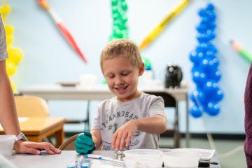 A young student smiles and wears a glove while working on a STEM project at a table, and teachers stand nearby
