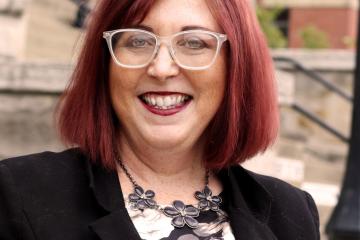 Female, smiling with shoulder length red hair, wearing glass, black and white blouse with a black blazer