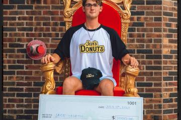 Jack Legath wears a Jeff's Donuts shirt and hat and sits on a throne after winning an eating competition