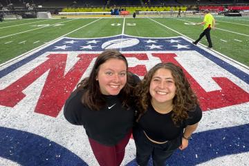 Two women stand on an NFL logo on a football field