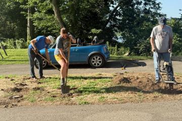 Three people work on outdoor garden space with shovels, rakes, tiller.