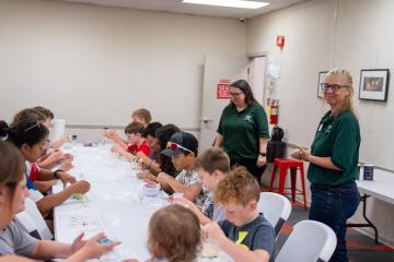 Local children work on projects at a long table while two OHIO representatives stand nearby to assist them at a Library STEM Day event