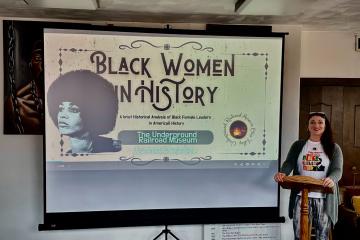 Photo of a female with long black hair standing behind a wooden podium, next to a projection screen that show a slide stating "Black Women in History"