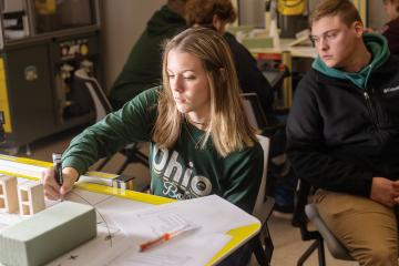 a female student in an Ohio University t-shirt creates a manufacturing model at a desk