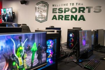 Computers in the foreground and a wall with "welcome to the esports arena" in the background. 
