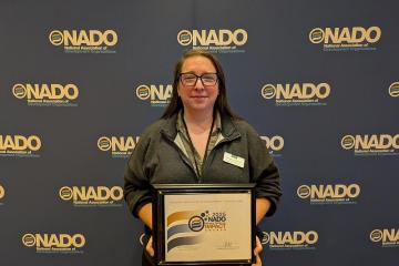 An OHIO representative holds the Impact Award while standing in front of a media wall that says NADO
