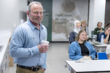 a man with a short gray beard and hair in a blue button-down shirt and khakis holds a coffee cup in front of a group of students sitting at desks