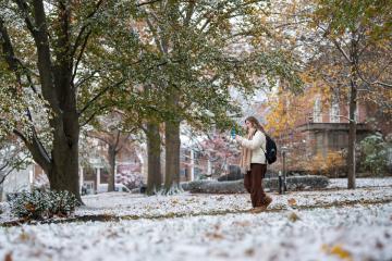 An OHIO student looks at her phone while walking on the College Green, which has light snow on the ground and colorful fall leaves on the trees