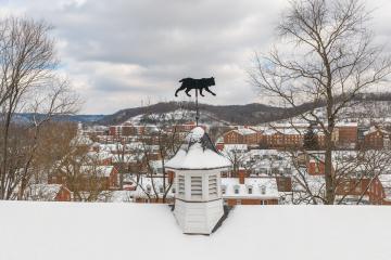 An image of a Bobcat weather vane on top of an Ohio University building, with many other buildings and the hillside in the background covered  in snow