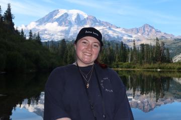 Jenna Pfeiffer stands in front of a large body of water with a mountain, tall trees and a blue sky in the background.