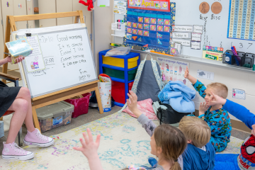 Student sits at front of classroom while students excitedly raise their hands
