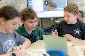 two female students drawing, one male teacher overlooking their work
