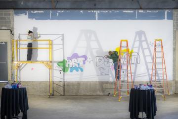 Two artists painting on the walls of Seigfred hall while its under construction