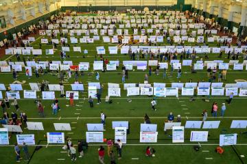 Rows of research posters, with students standing near them, are shown in the Walter Fieldhouse for the Student Research Expo.
