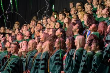 A large group of Heritage College graduates in green doctoral regalia look up and smile as green and silver streamers fall over them during their commencement ceremony.
